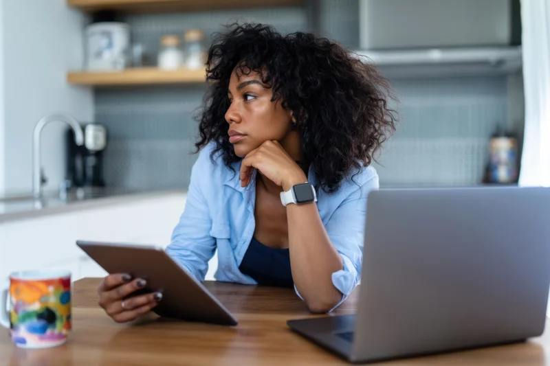 Woman sitting at wooden table in modern kitchen using tablet and laptop, thinking with hand on chin