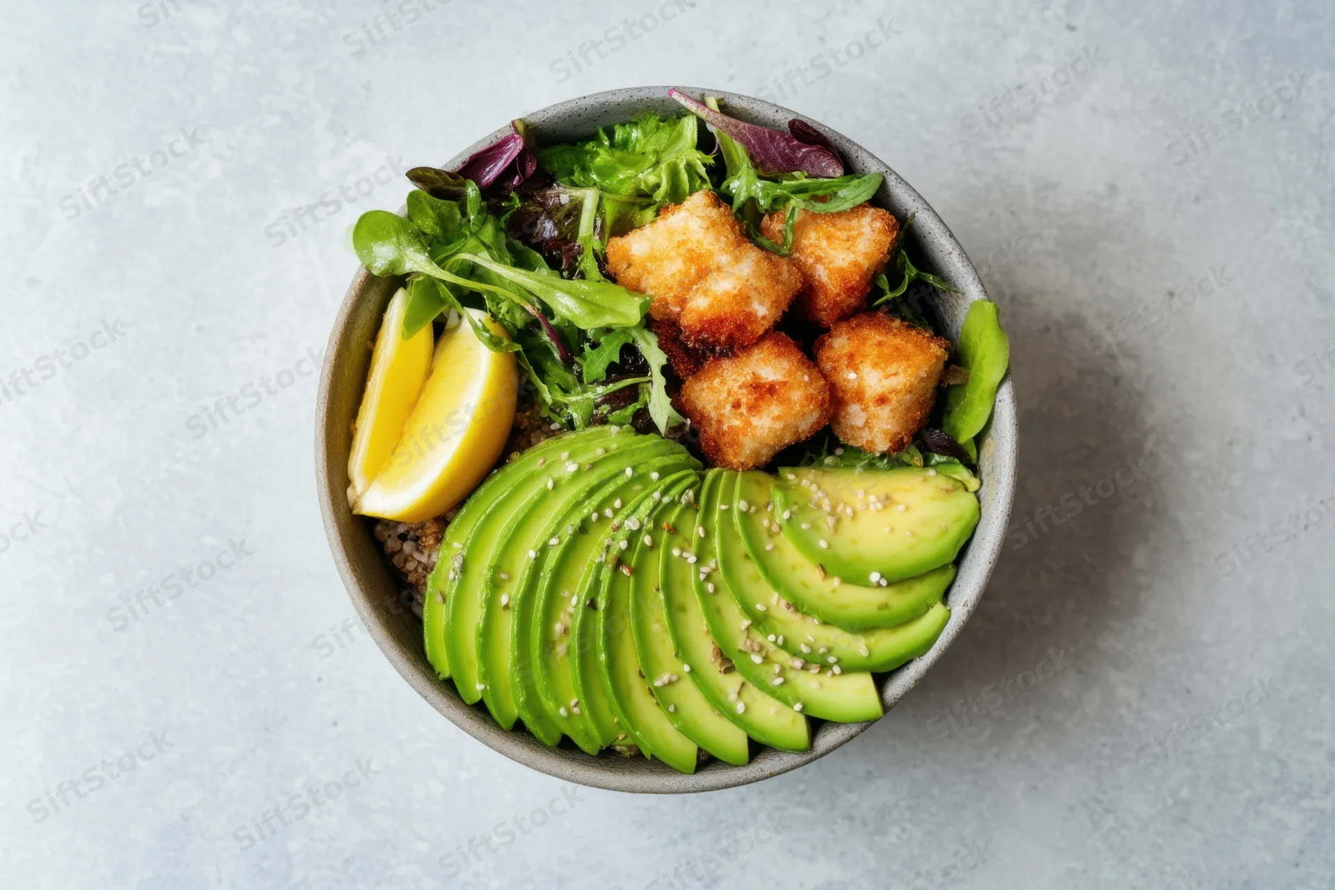 A salad bowl with fan-sliced avocado, crispy tofu, greens and lemon on a gray surface