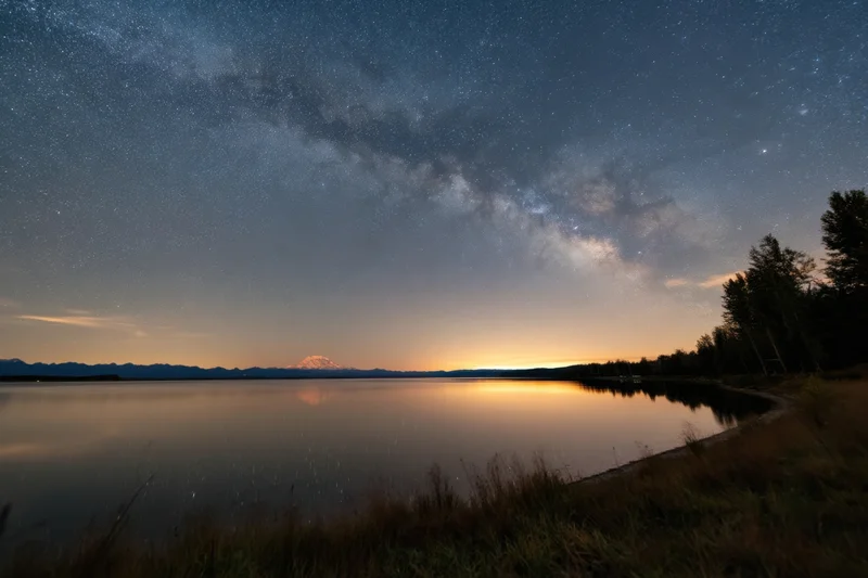 Star-filled night sky over a calm lake reflecting distant mountains and twilight horizon