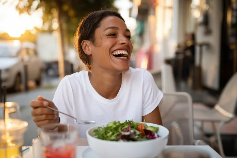 Woman laughing while holding fork over salad bowl at outdoor cafe table
