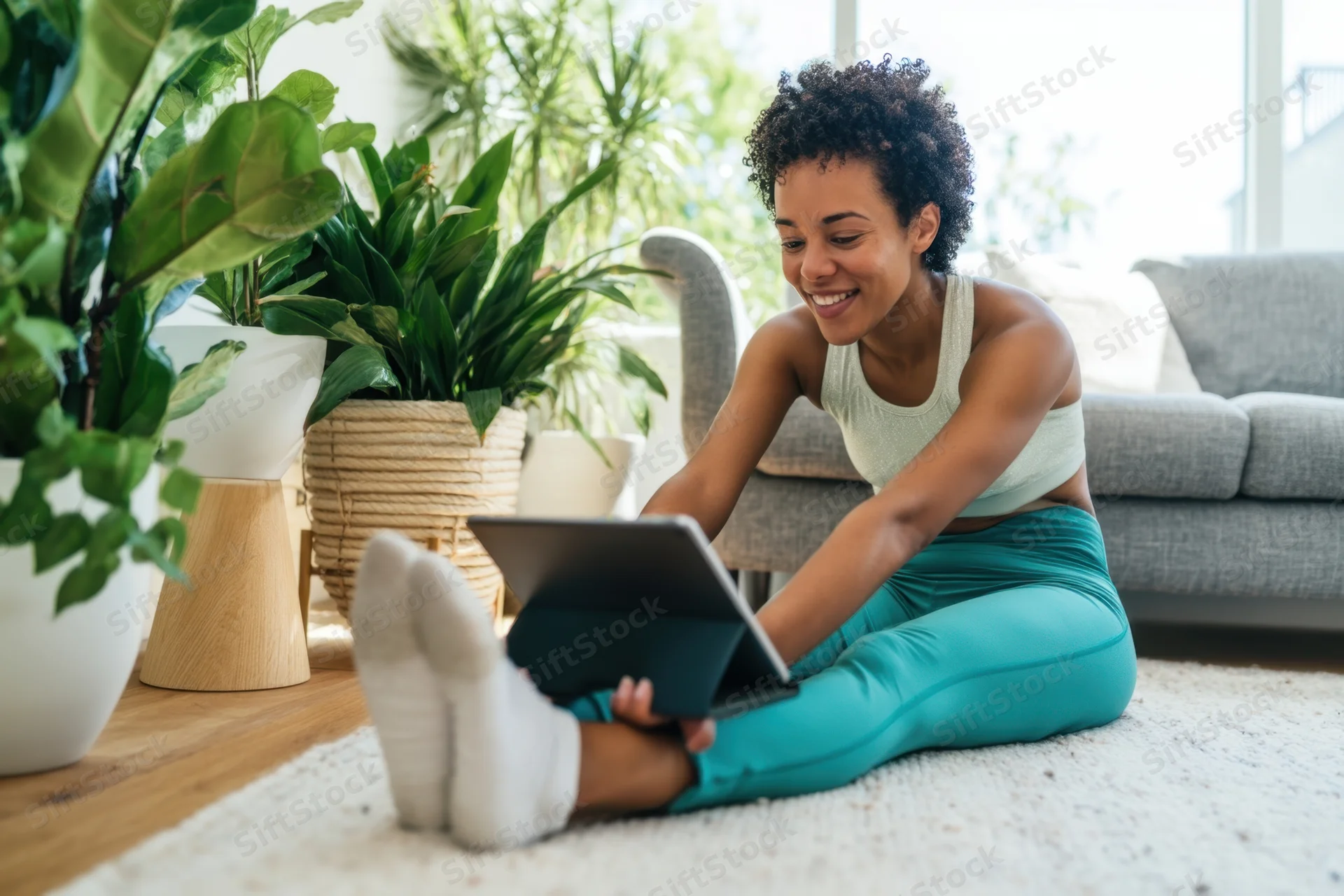 Woman sitting on floor using tablet in living room with plants around her