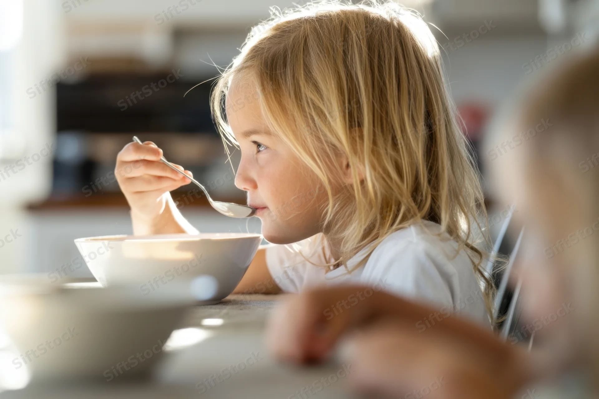 Young blonde girl eating cereal from a white bowl at a kitchen table in the morning