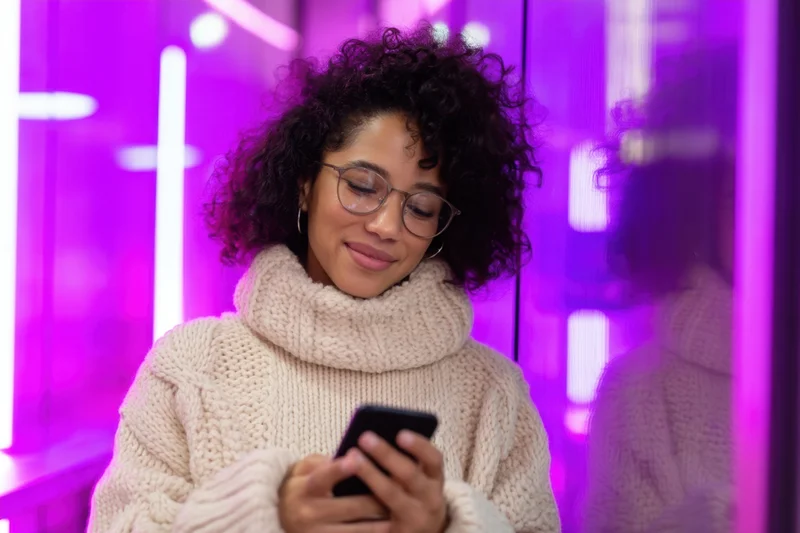 Young woman smiling while using smartphone in neon-lit urban interior
