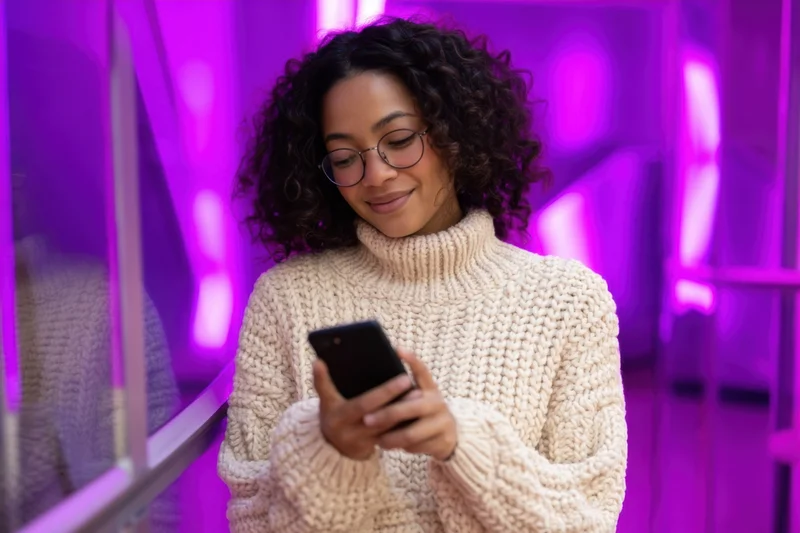 Young woman with curly hair and glasses smiling while using smartphone in a purple neon-lit hallway
