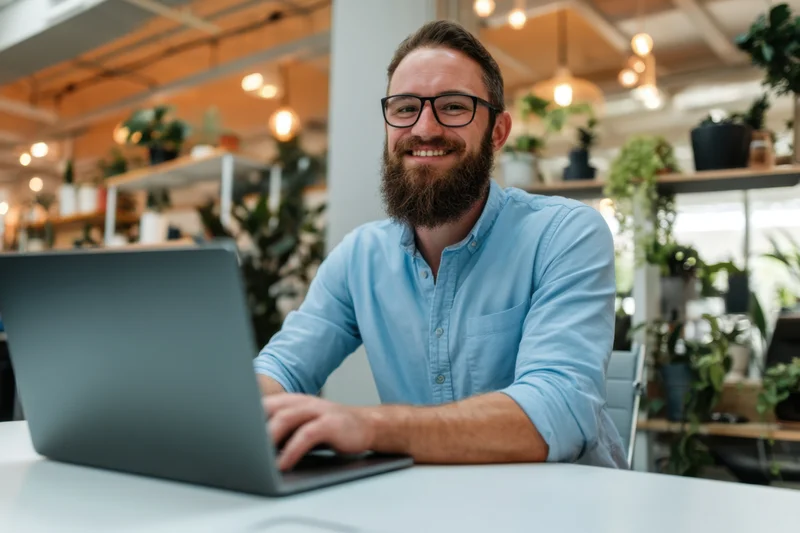 Bearded man smiling while using laptop at white desk in modern office with plants and warm lighting