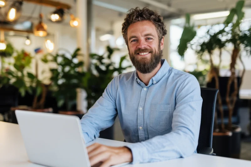 Bearded man smiling while using laptop at white desk in modern office with plants and pendant lights