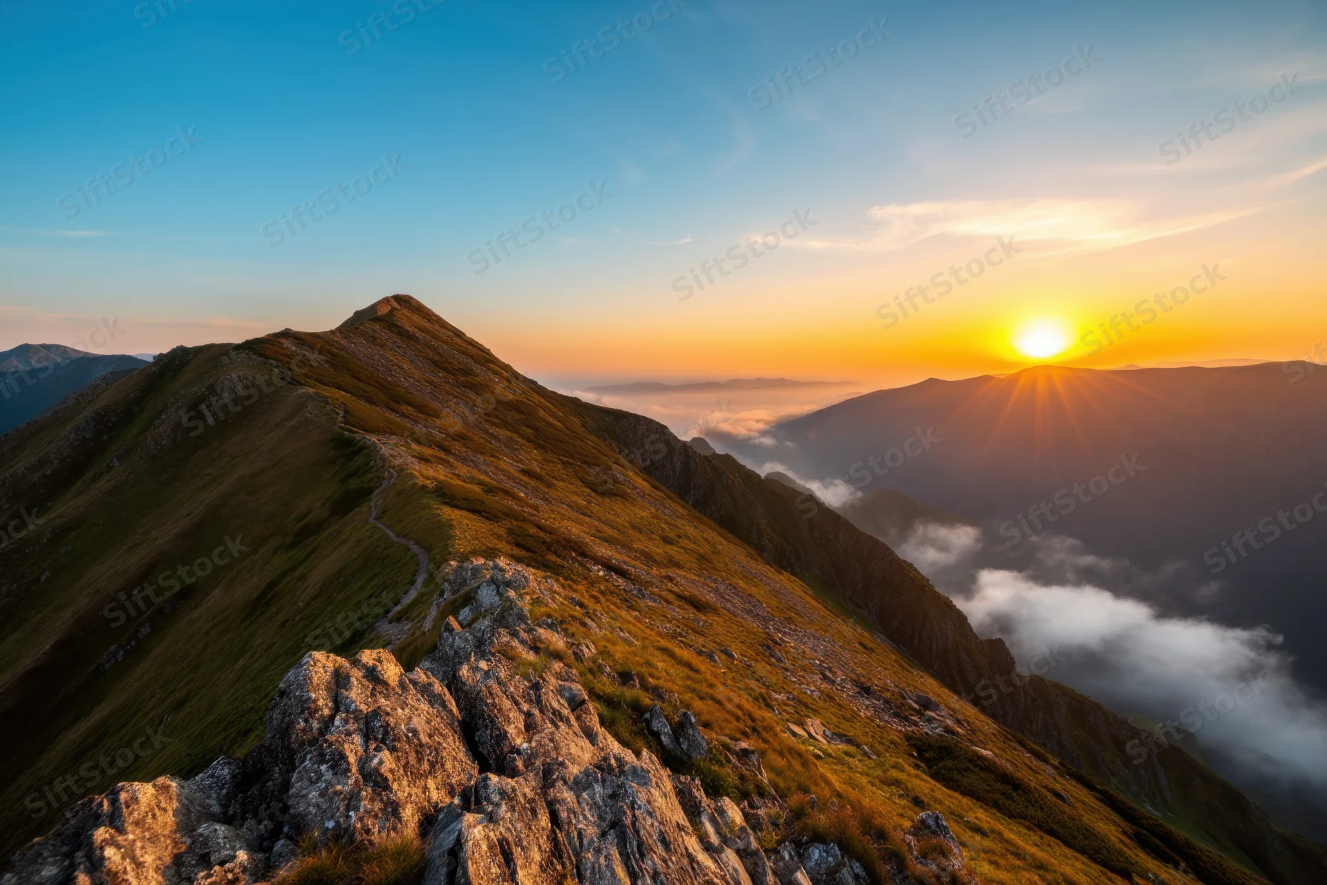 Mountain peak at sunset with clouds below and a winding trail along the ridge