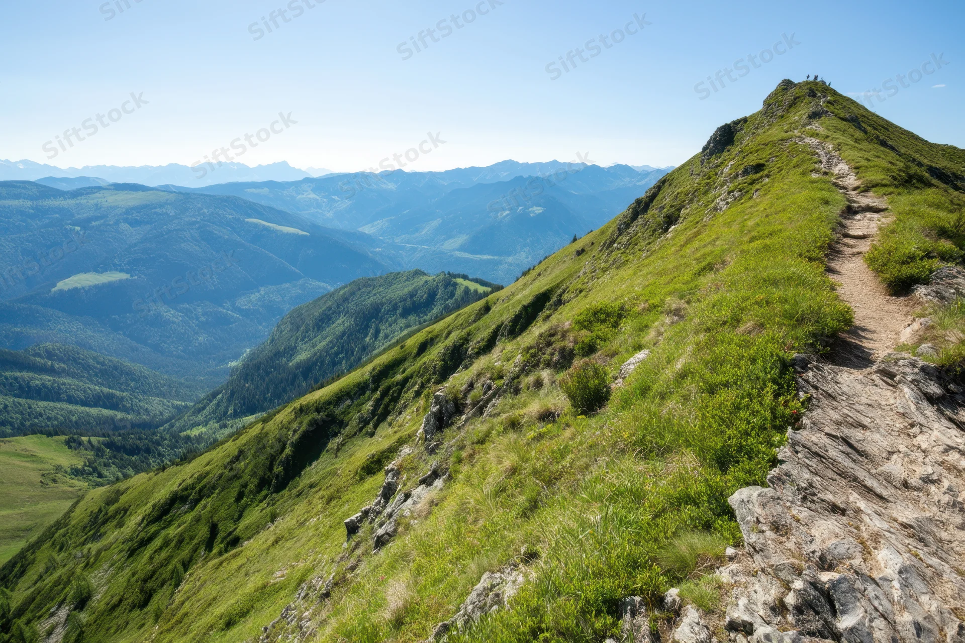 Narrow dirt trail winding along grassy mountain ridge overlooking valley and distant peaks