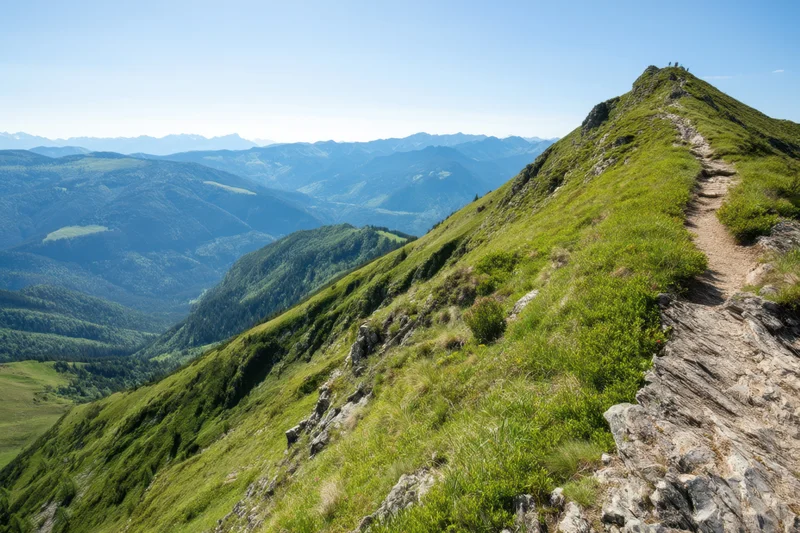Narrow dirt trail winding along grassy mountain ridge overlooking valley and distant peaks