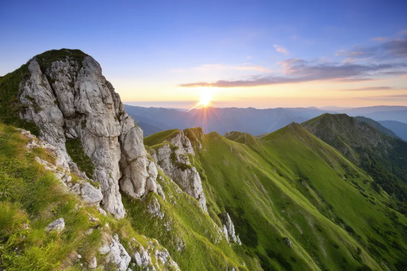 Mountain peak at sunrise overlooking a green valley with rocky ridges and distant peaks under a clear sky