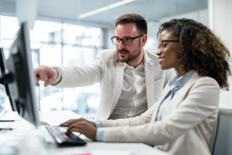 Two colleagues in business attire collaborating at a desk by a computer monitor
