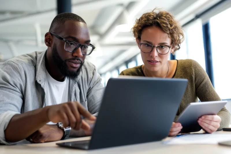 Two coworkers collaborating on a laptop and tablet in a modern office
