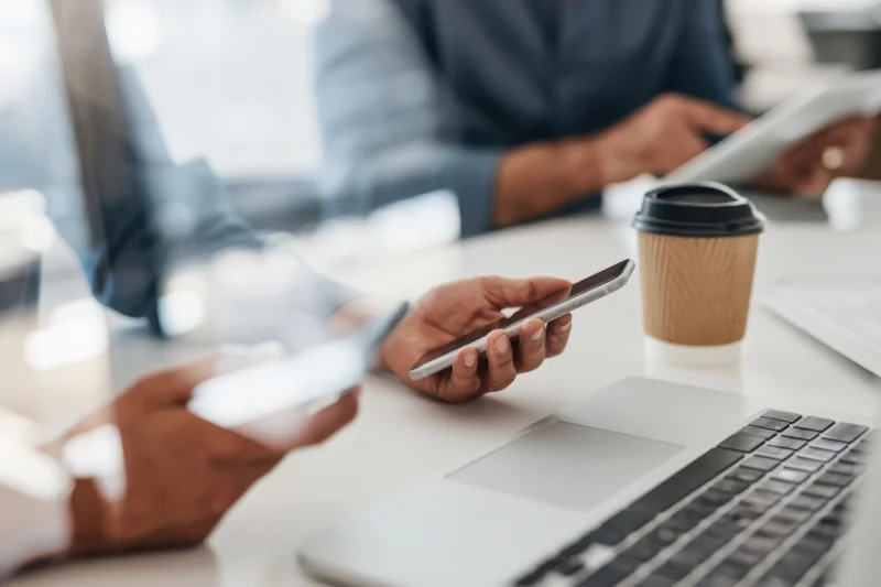 Hands using smartphone at office desk with laptop and coffee during work meeting