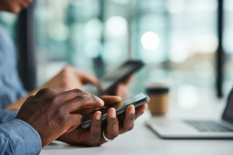 Hands typing on a smartphone at a desk in an office with a laptop and coffee nearby