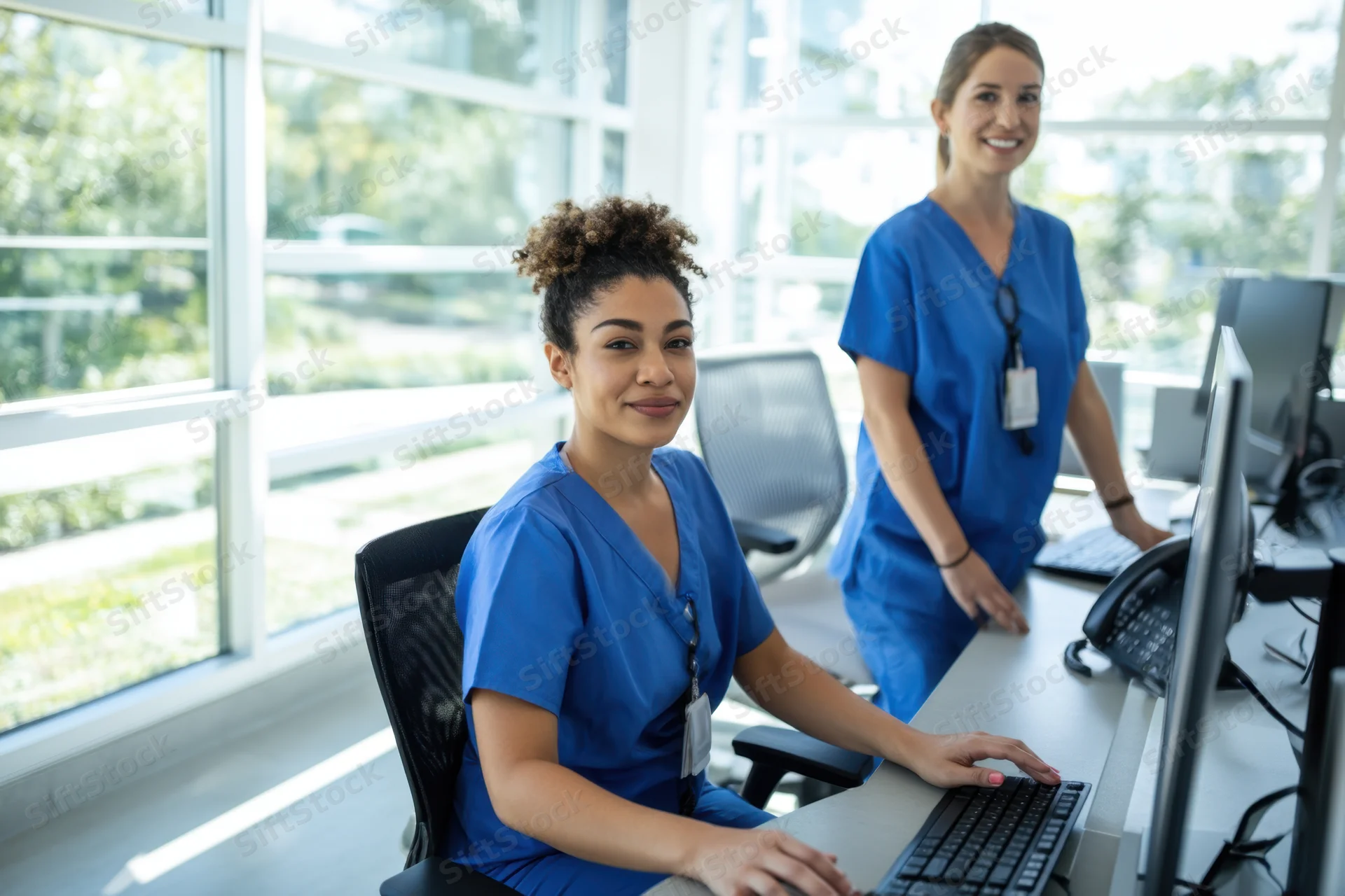 Two nurses in blue scrubs working at computers in a modern office with large windows