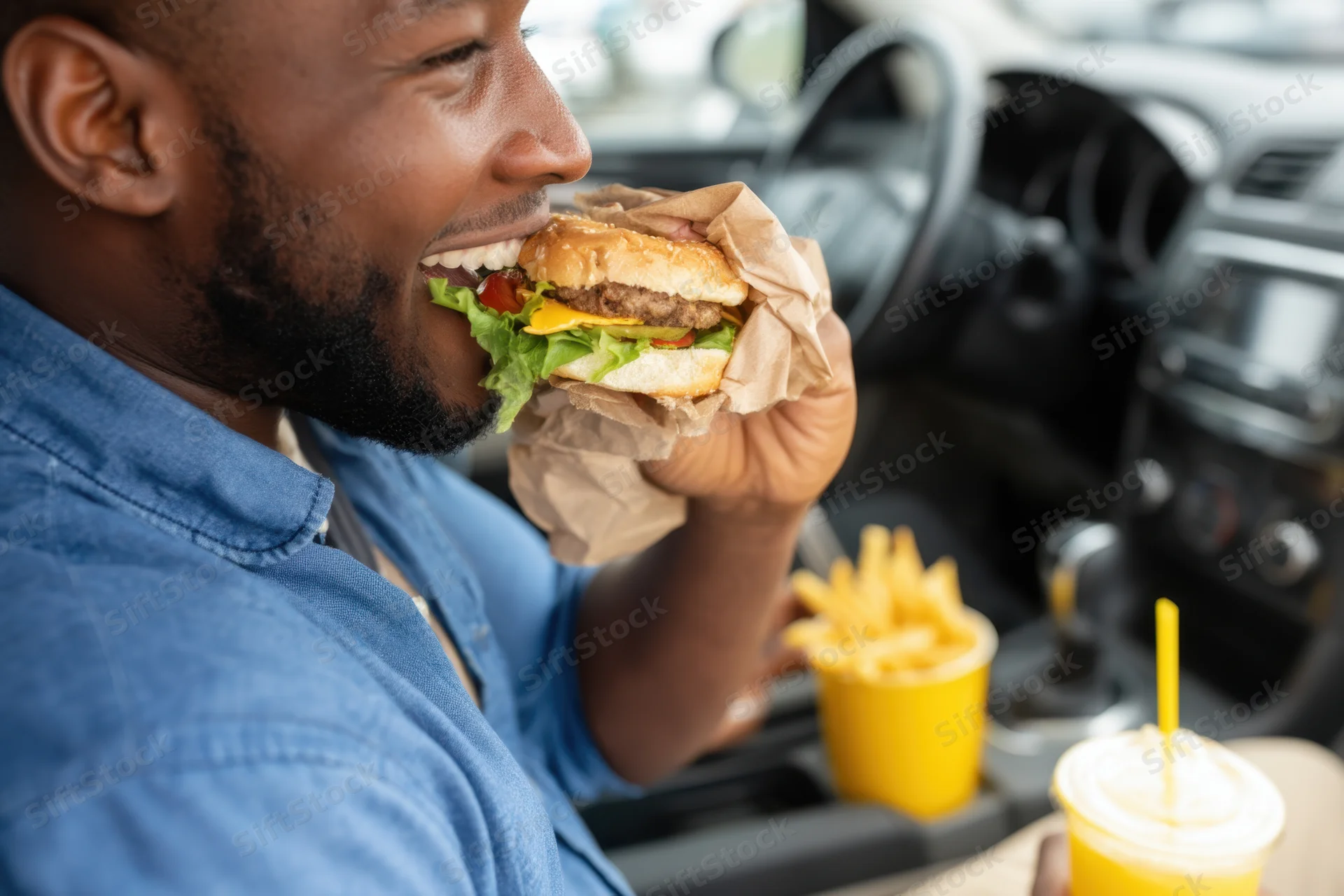 Man eating a cheeseburger in a car with fries and soda on the console