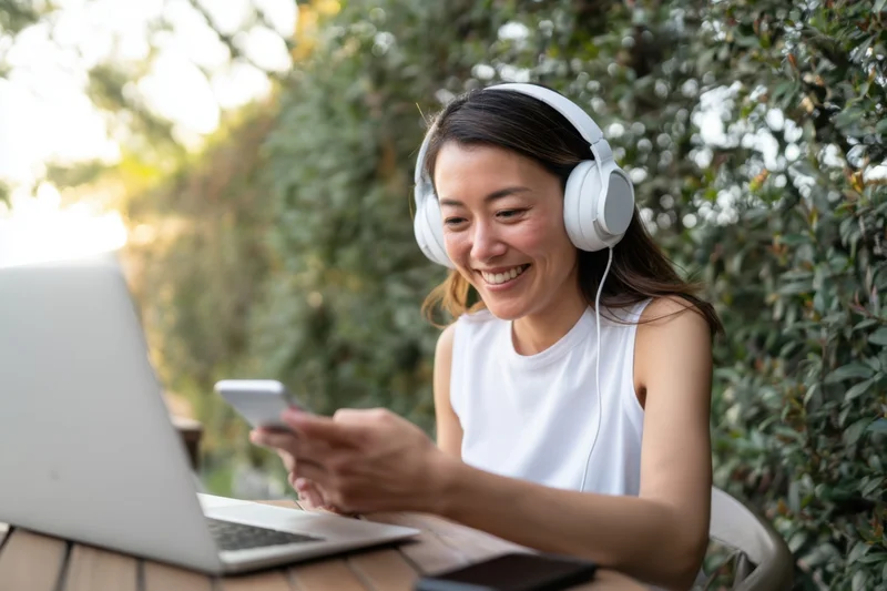 Woman smiling while using smartphone and laptop with headphones at outdoor table in garden