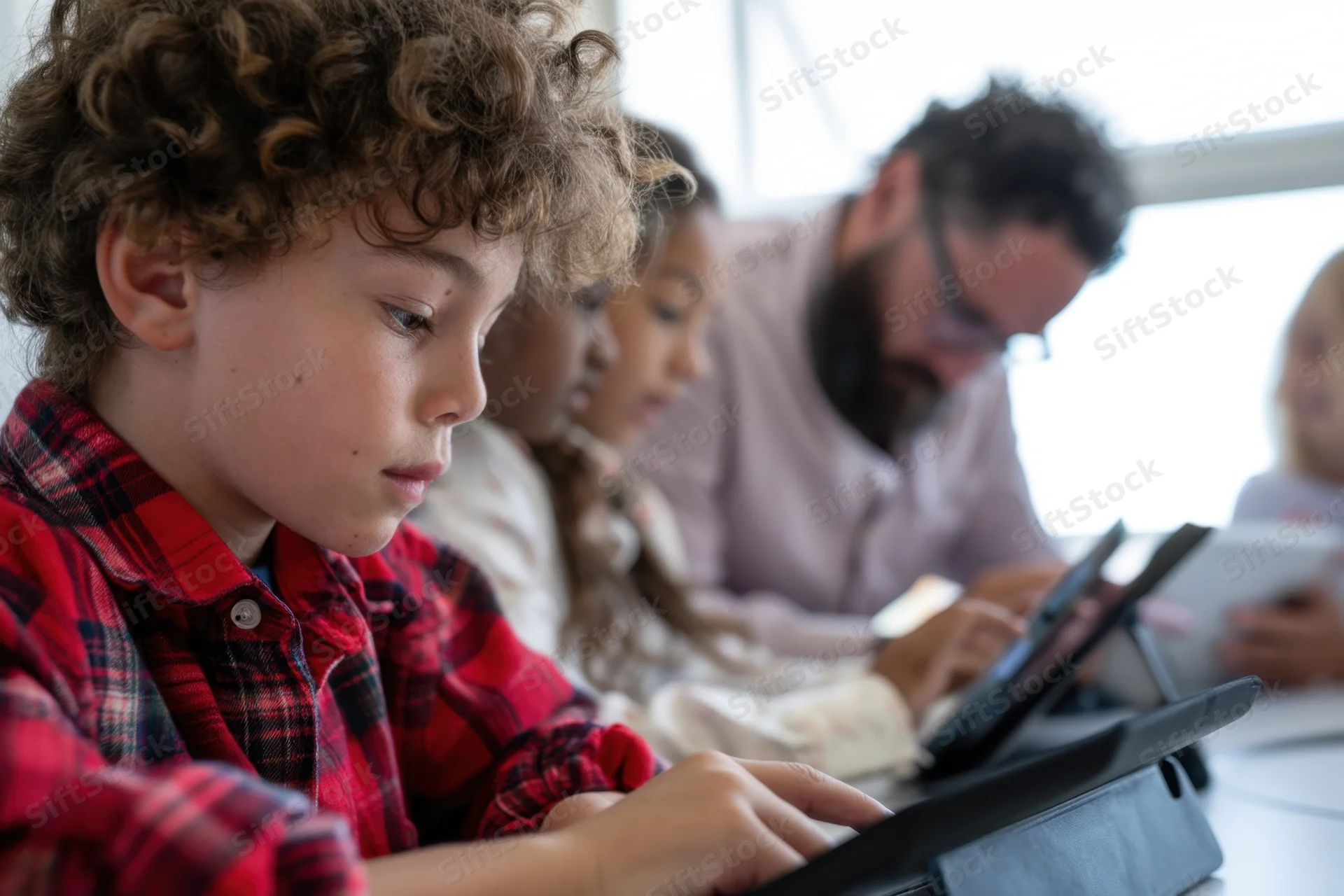 Young boy in red plaid shirt concentrating on tablet in classroom with teacher and peers nearby