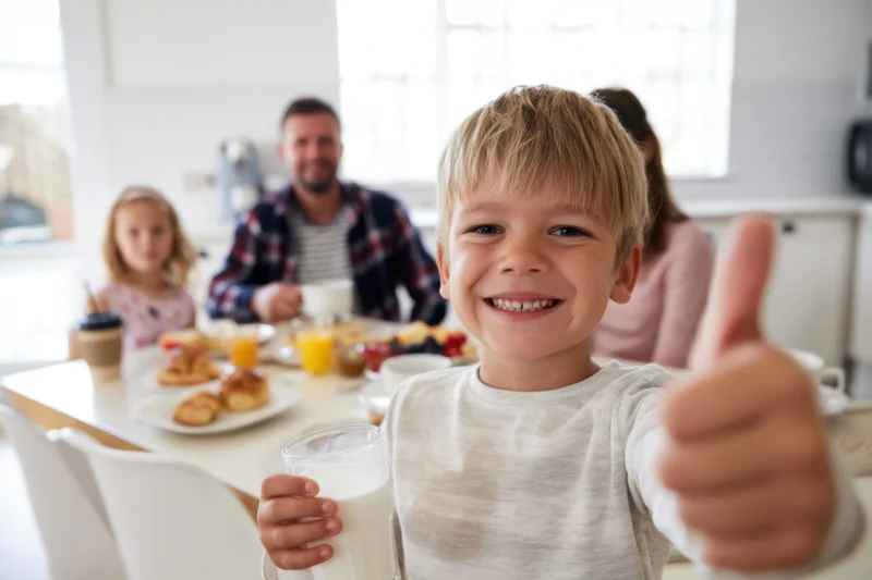 Smiling boy giving thumbs up while holding a glass of milk at breakfast table with family in kitchen