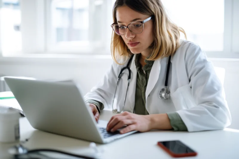 Doctor in white coat using laptop at desk in office