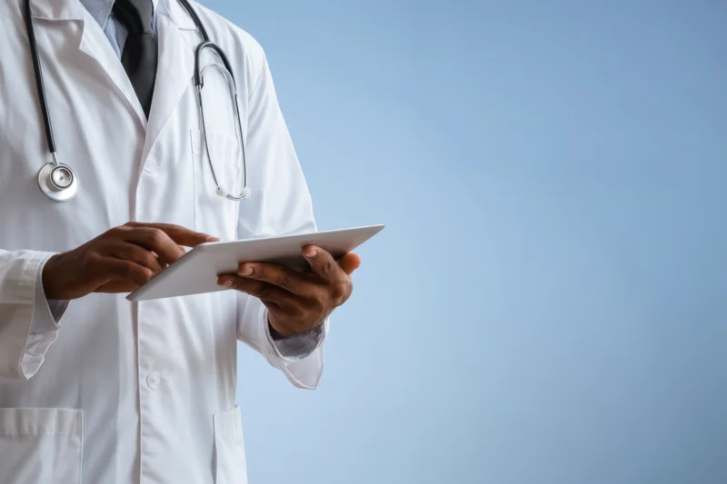 Doctor in white coat using tablet with stethoscope around neck against blue background