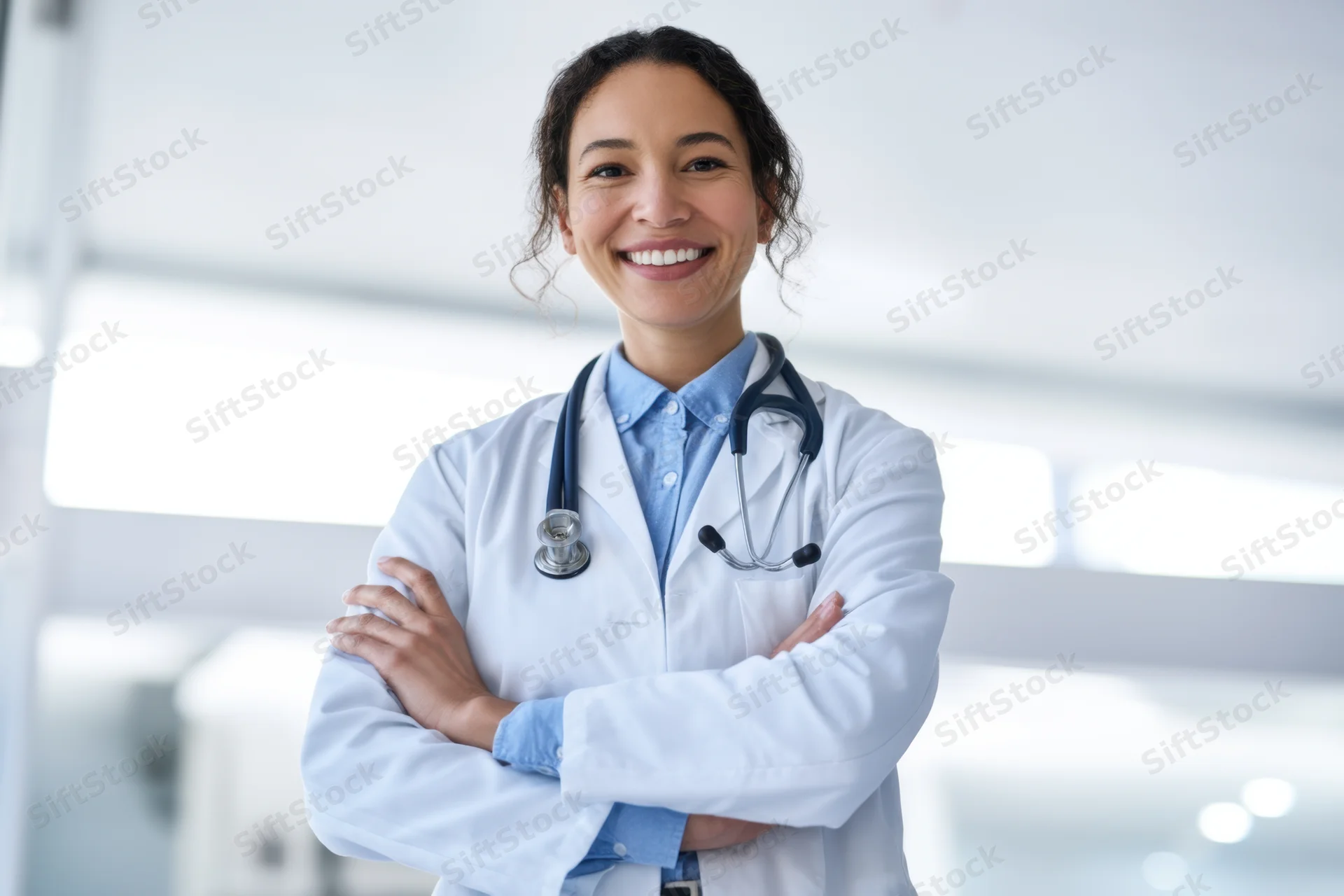 Female doctor in white lab coat with stethoscope smiling with arms crossed in clinic