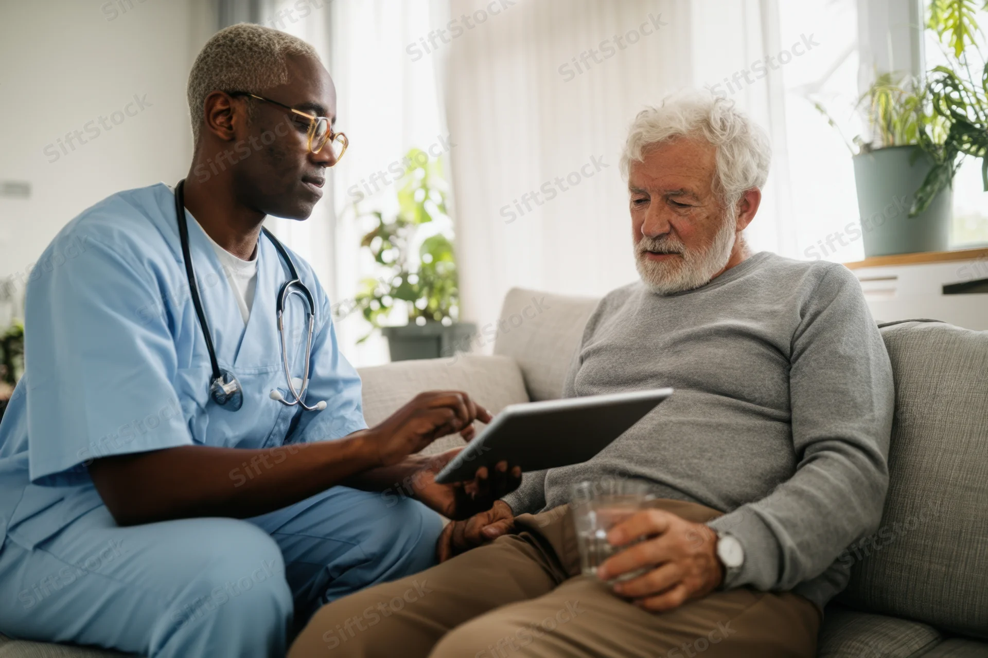 Doctor in light blue scrubs showing tablet to senior patient during home visit on couch