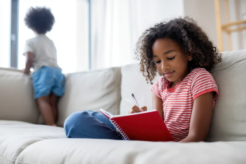 Girl writing in a red notebook while sitting on a light sofa at home