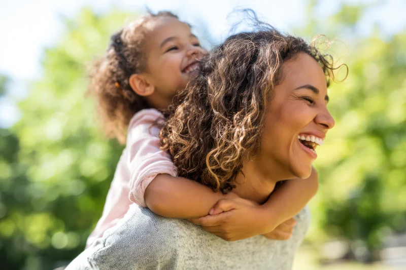 Woman laughing with child on her back in a sunlit park