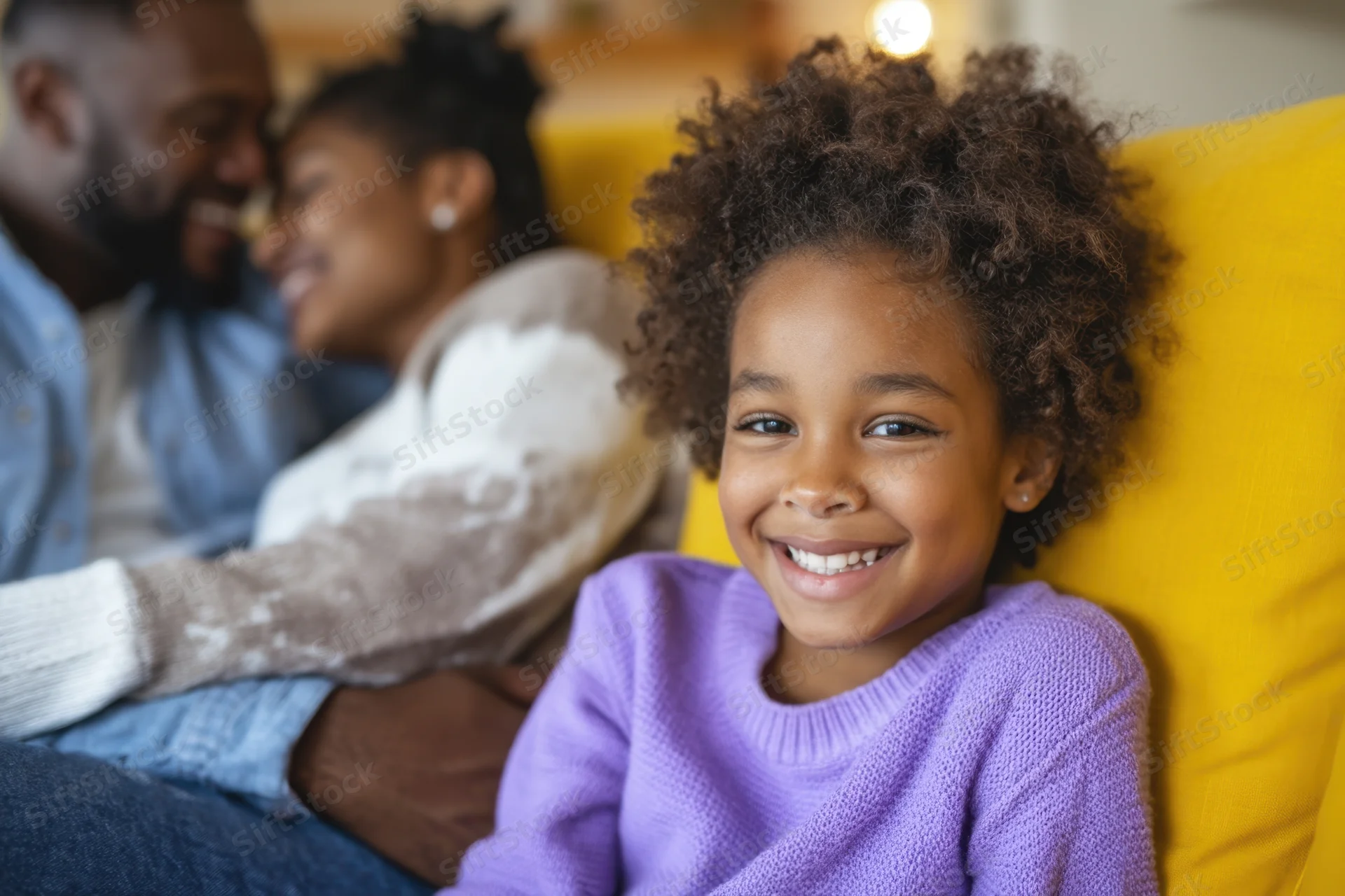 Young girl smiling on a yellow couch with parents embracing behind her in a living room