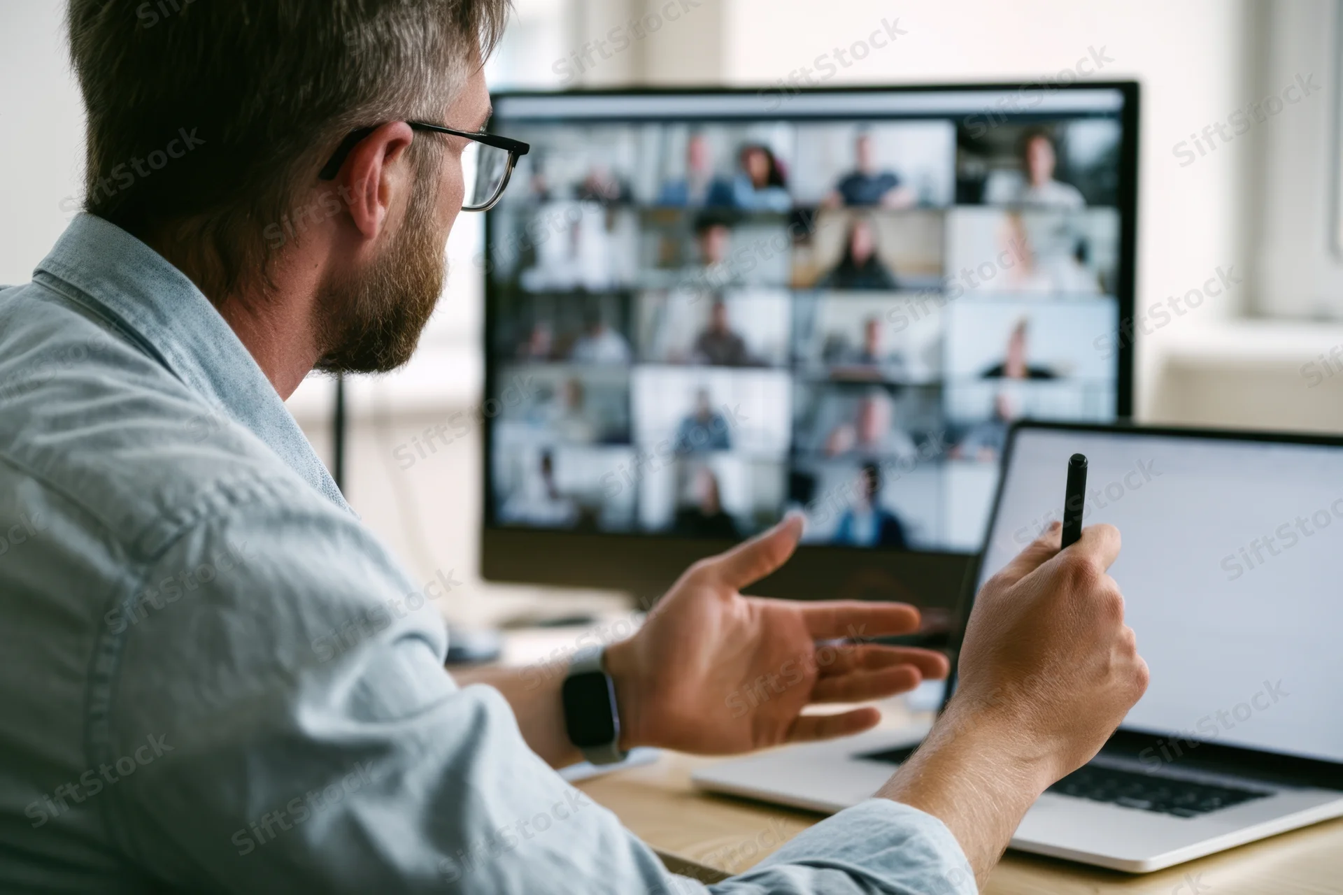 Man in casual shirt and glasses gesturing during video conference on laptop and monitor in office workspace