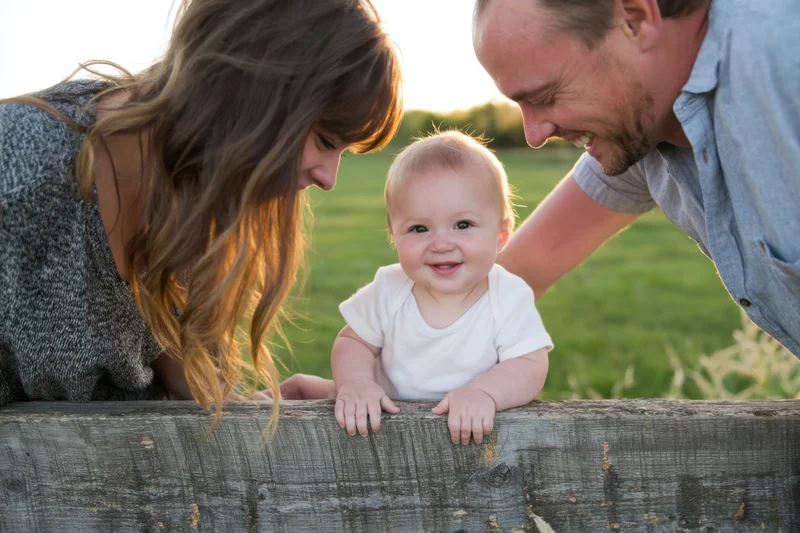 Smiling baby perched on a weathered wooden fence with parents leaning in from both sides in a grassy field