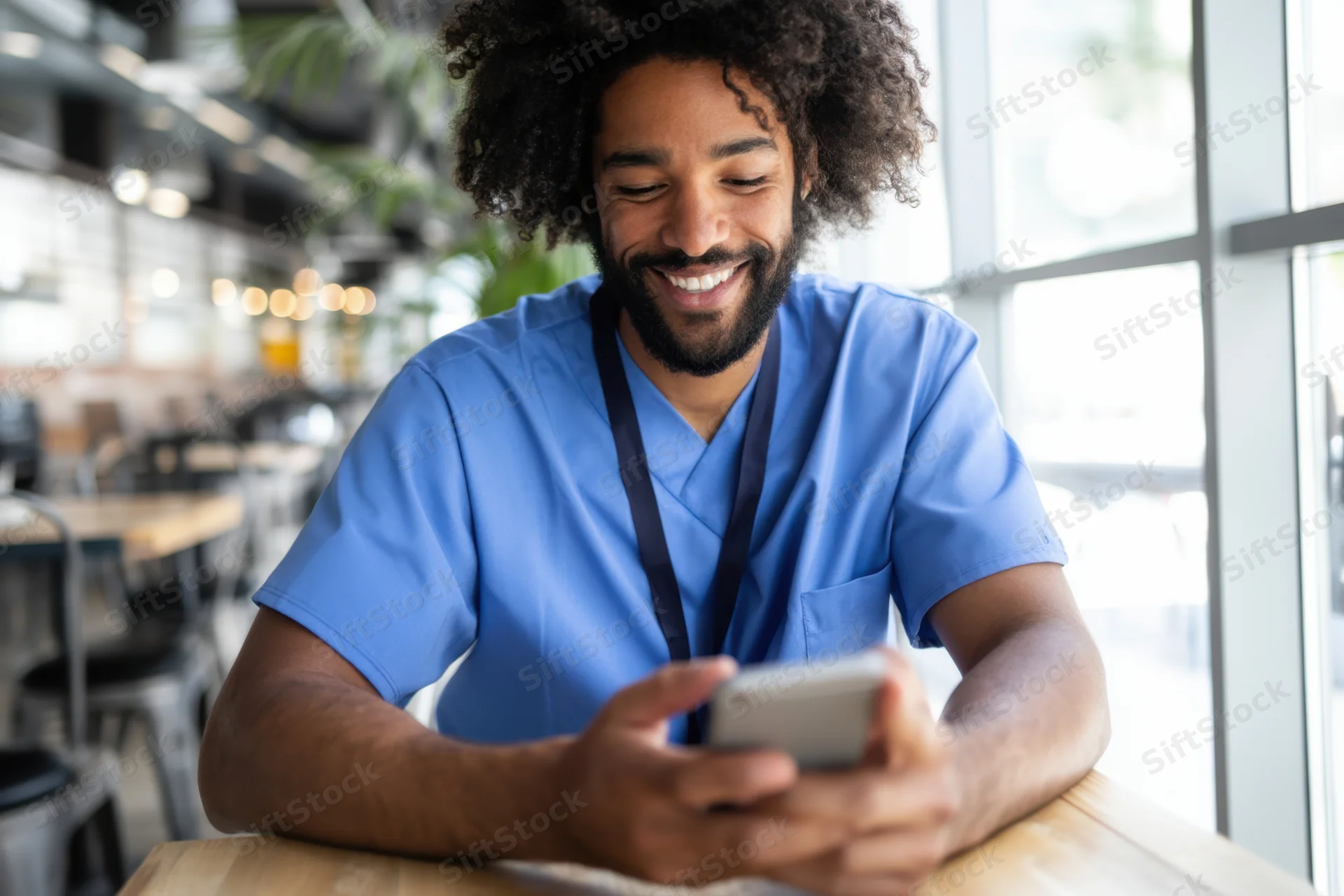 Man in blue scrubs smiling while using smartphone at cafe table