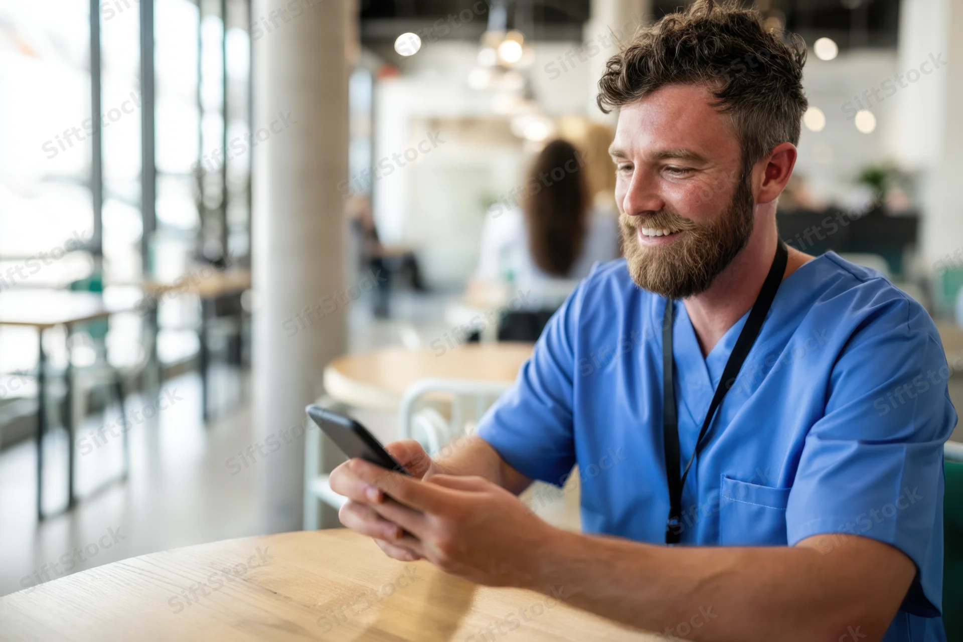 Bearded man in blue scrubs smiling while using smartphone at cafe table