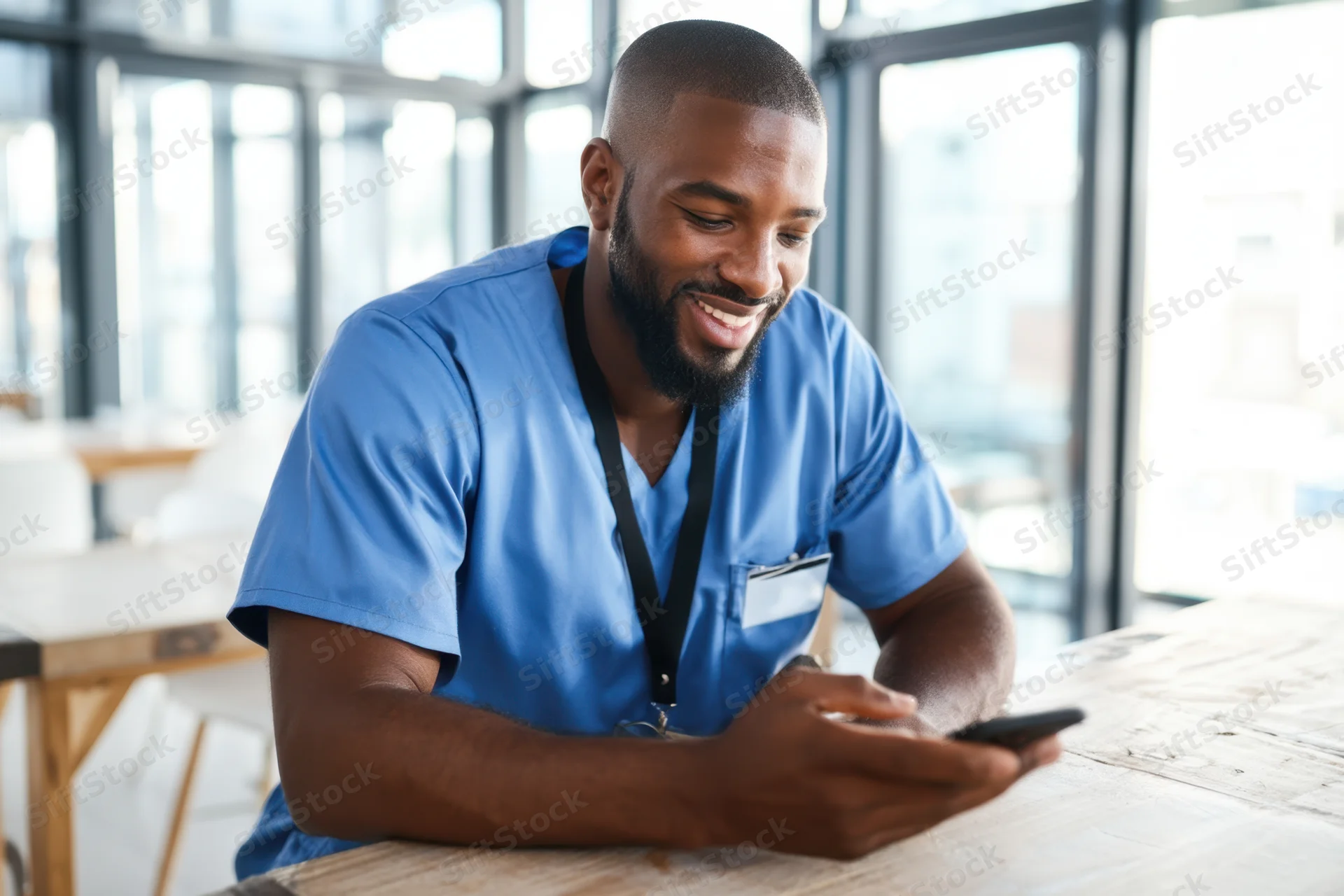 Smiling bearded man in blue scrubs using smartphone at table in bright office