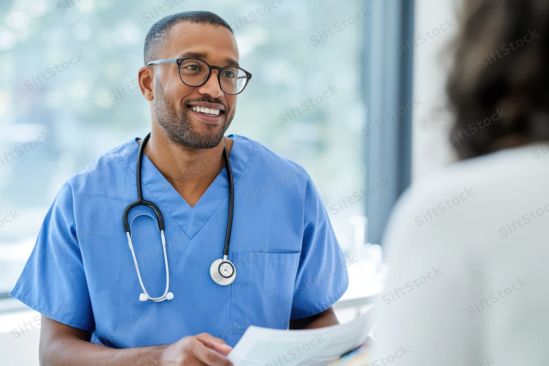 Smiling male doctor in blue scrubs holding papers during patient consultation in clinic