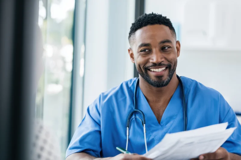 Smiling male healthcare professional in blue scrubs holding paperwork during patient consultation in clinic