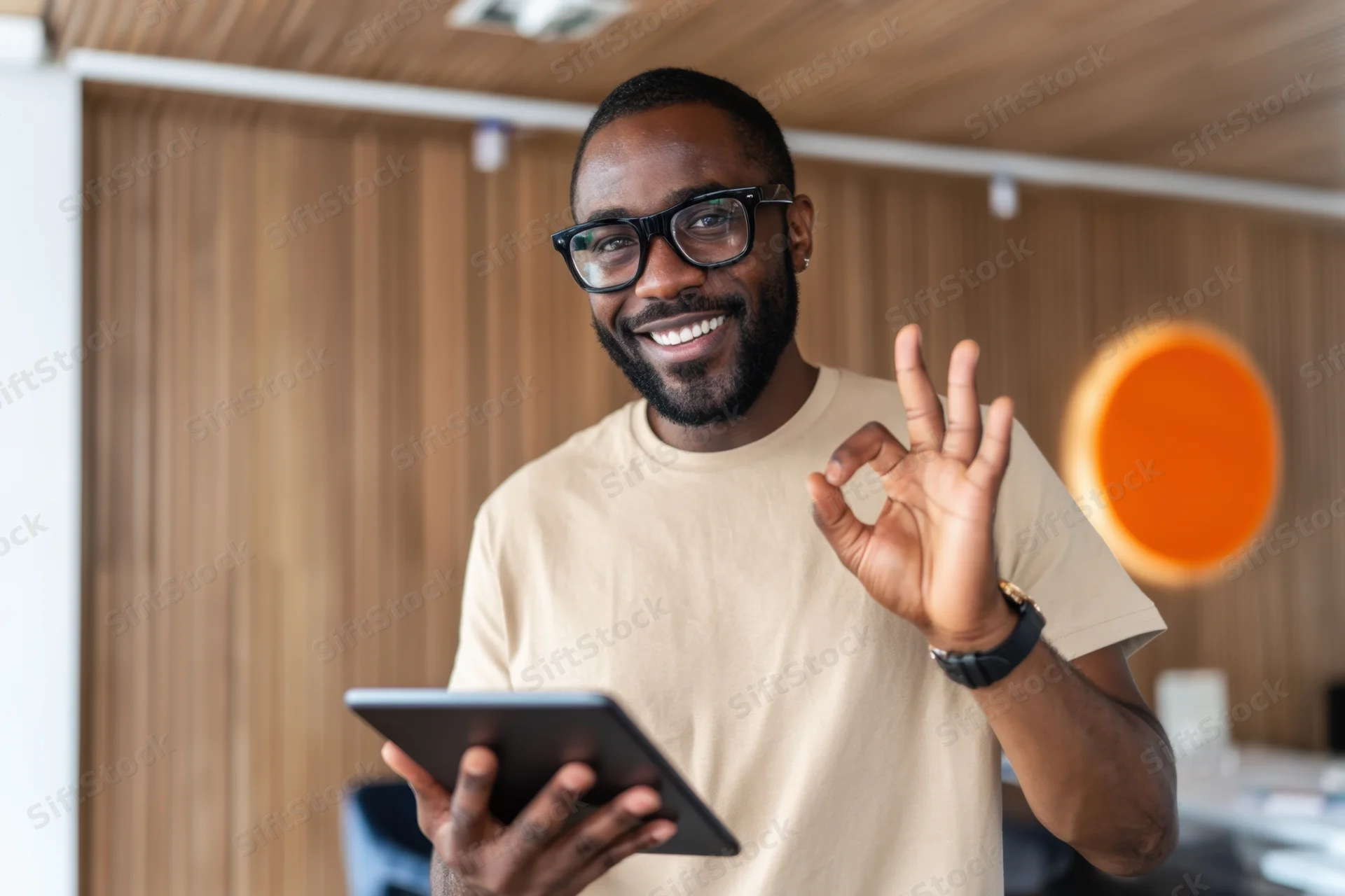 Smiling man in beige t-shirt and black glasses holding tablet and making ok gesture in modern office workspace