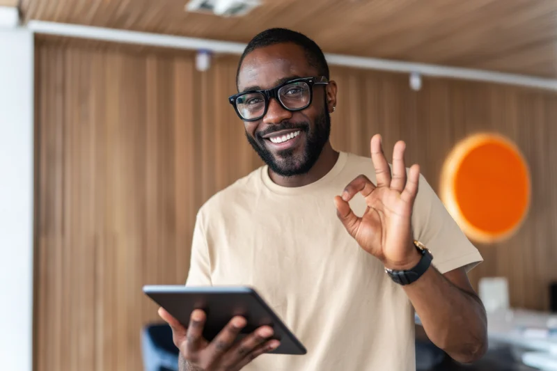 Smiling man in beige t-shirt and black glasses holding tablet and making ok gesture in modern office workspace