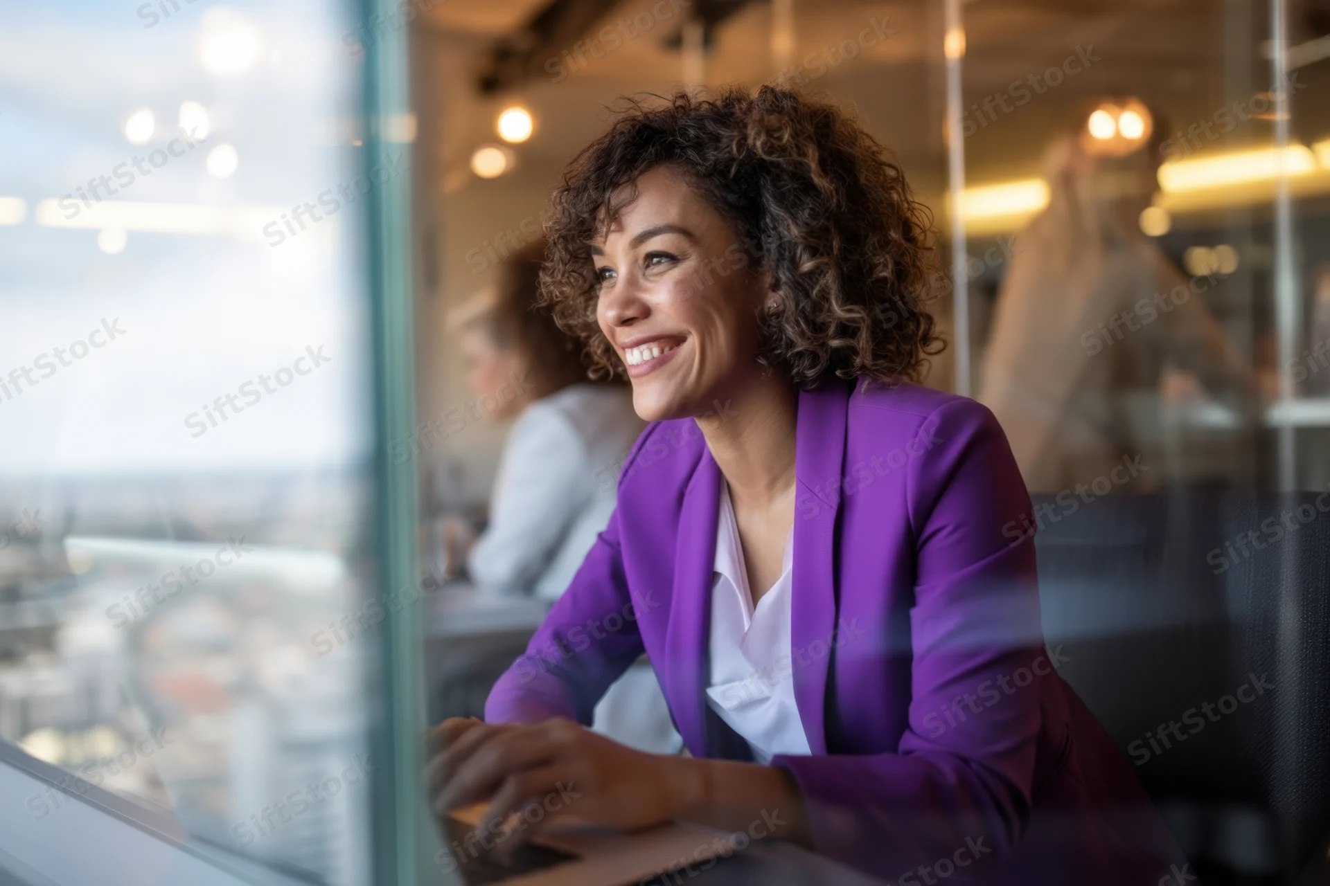 Woman in purple blazer smiling while working on laptop at glass-walled office desk overlooking city