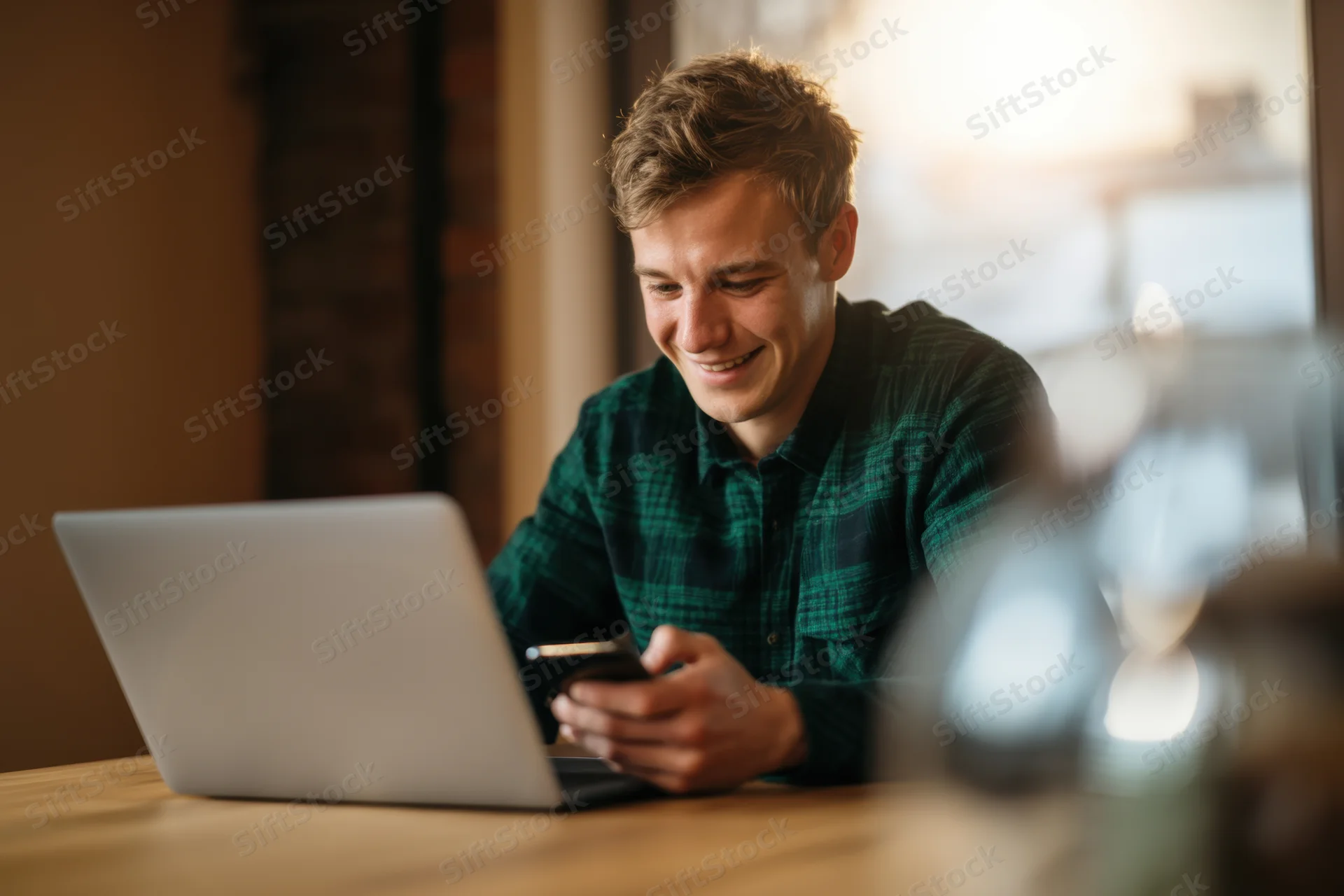 Young man smiling while using laptop and smartphone at wooden desk indoors