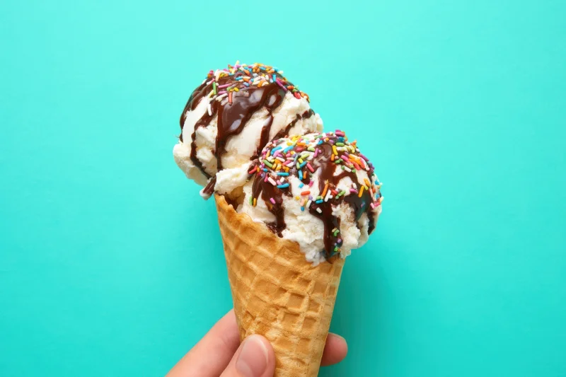 Hand holding a waffle cone with two scoops of vanilla ice cream topped with chocolate syrup and colorful sprinkles against a turquoise background
