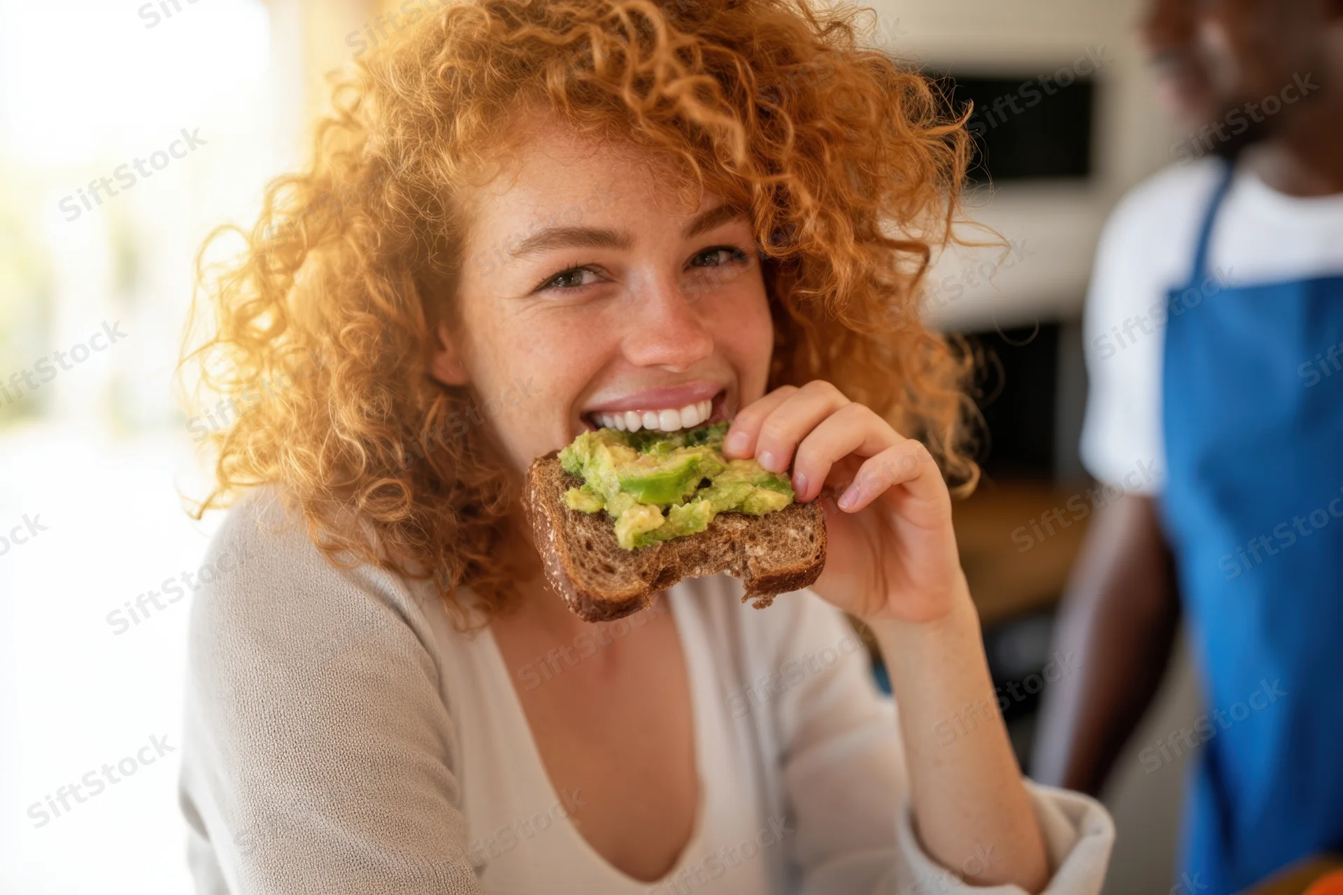 Woman biting into avocado toast in a kitchen while smiling