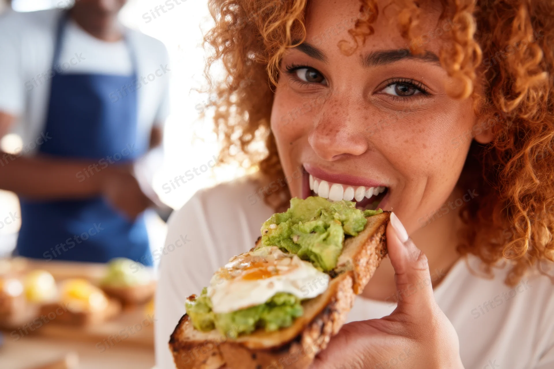 Woman biting into avocado toast with egg in a kitchen setting