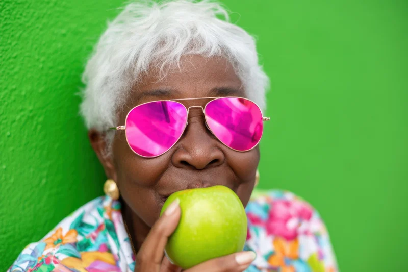 Senior woman with short white hair wearing pink mirrored sunglasses eating a green apple against a green wall