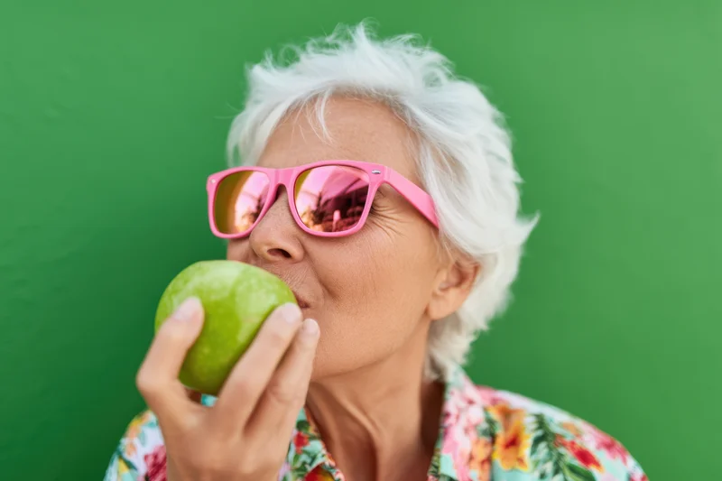 Senior woman with white hair wearing pink sunglasses eating a green apple against a green background