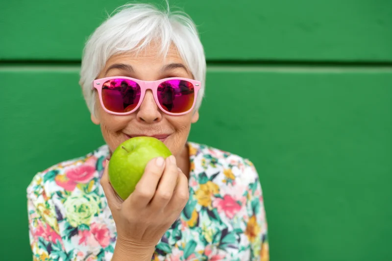 Senior woman smiling while holding a green apple against a green wall