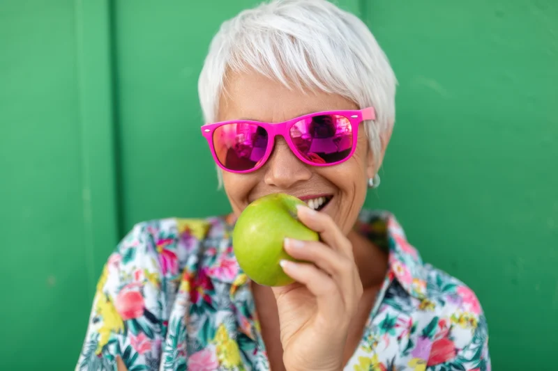 Senior woman smiling while eating a green apple outdoors wearing pink mirrored sunglasses and a floral shirt