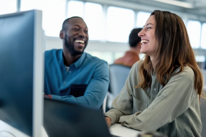 Two coworkers laughing together at a desk in a bright office