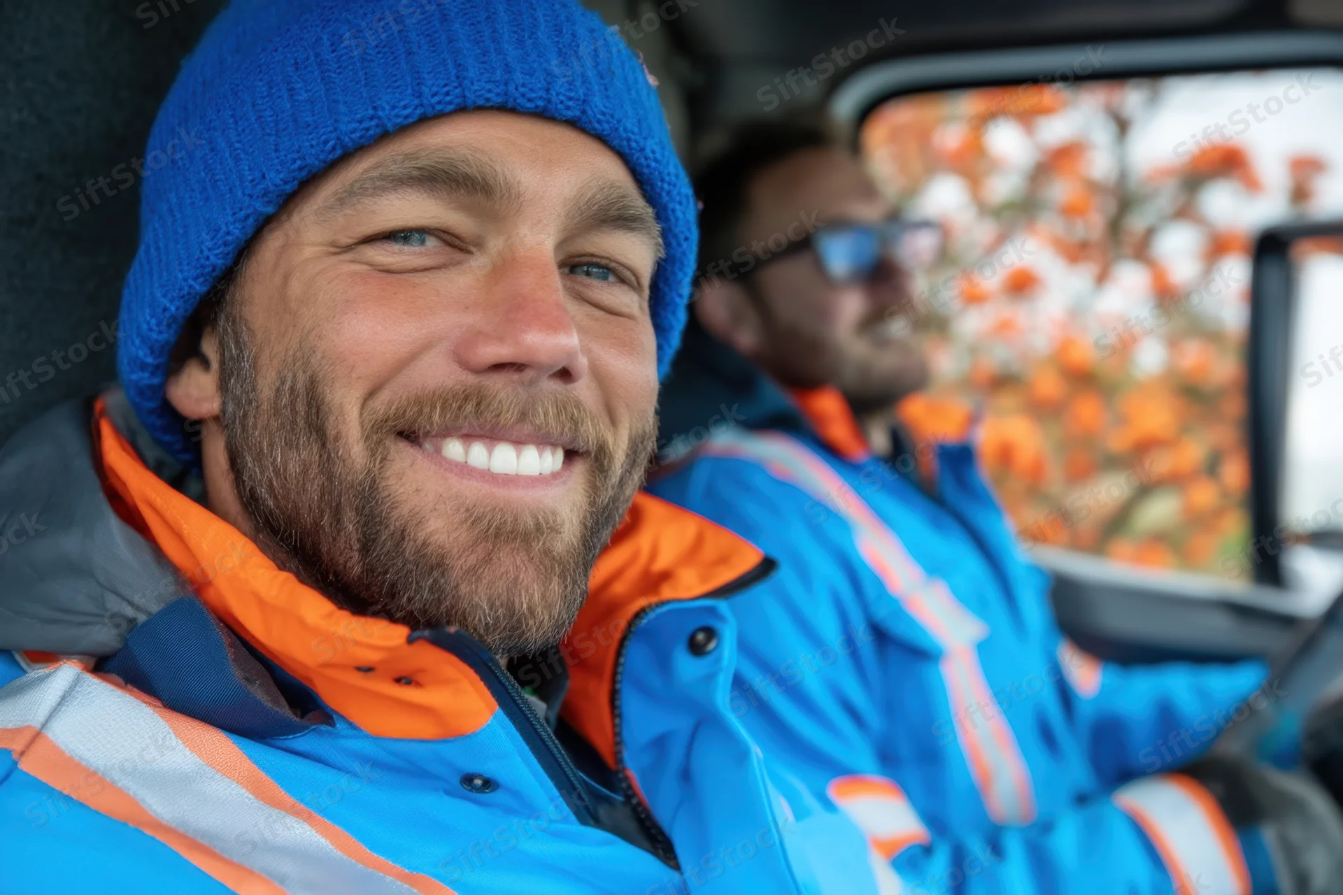Two men in blue and orange high-visibility jackets smiling inside a car during autumn